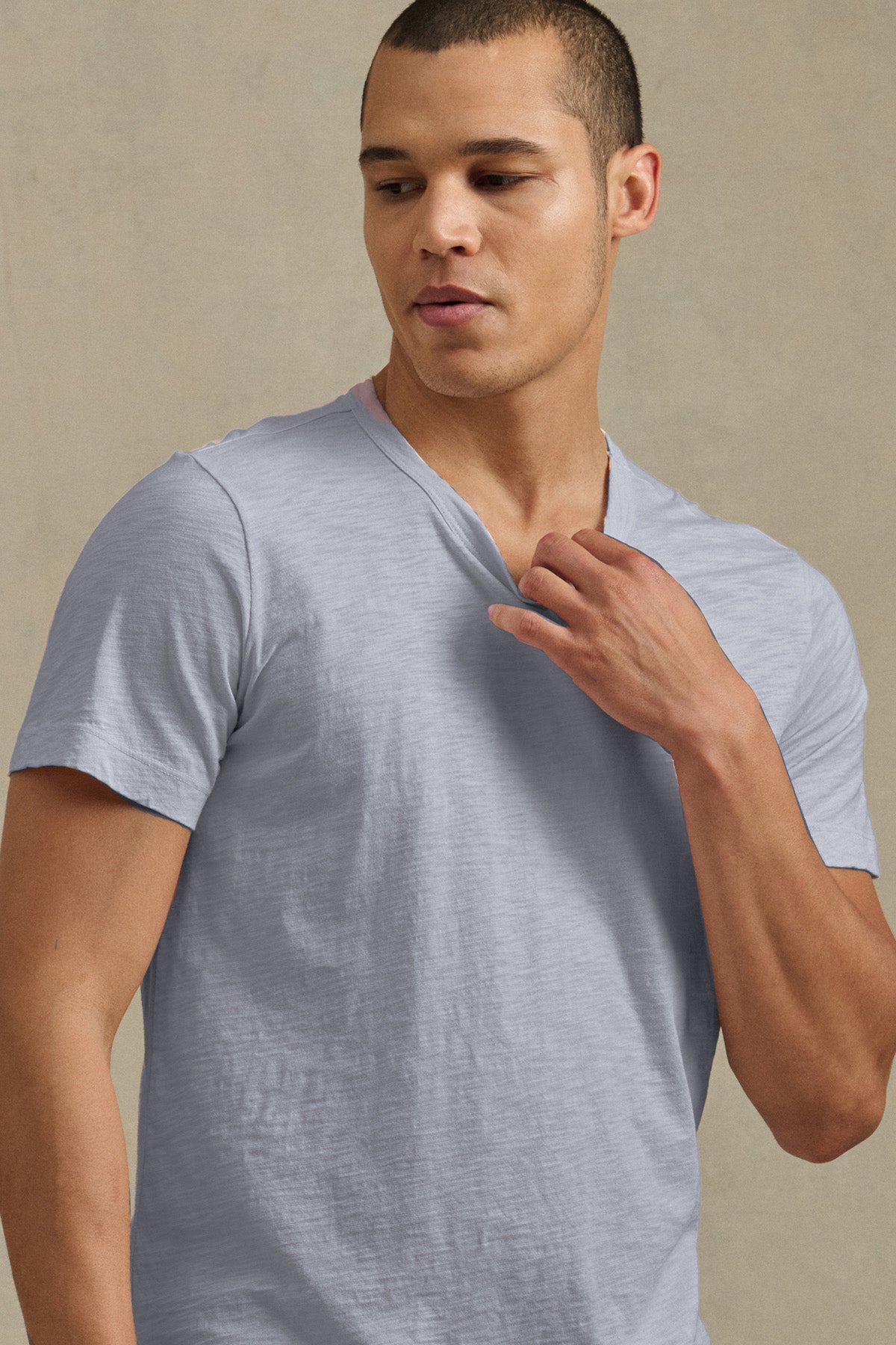 A man wearing the light gray AMARO TEE by Velvet by Graham & Spencer stands against a neutral background, looking down and to the side while holding his collar.-41428277526721