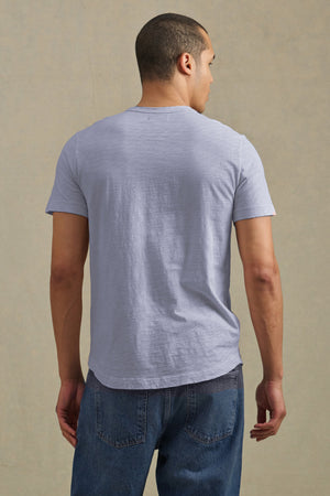 A man faces away from the camera, wearing the light gray AMARO TEE by Velvet by Graham & Spencer with a classic crew neck and blue jeans against a beige background—a versatile casual layering piece.