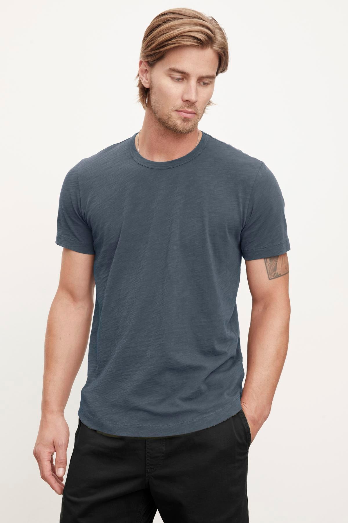   A person with long hair wearing a relaxed stylish feel, casual, short-sleeved dark gray Velvet by Graham & Spencer AMARO TEE and black pants stands against a plain light background. They have a neutral expression and a tattoo on their left arm. 