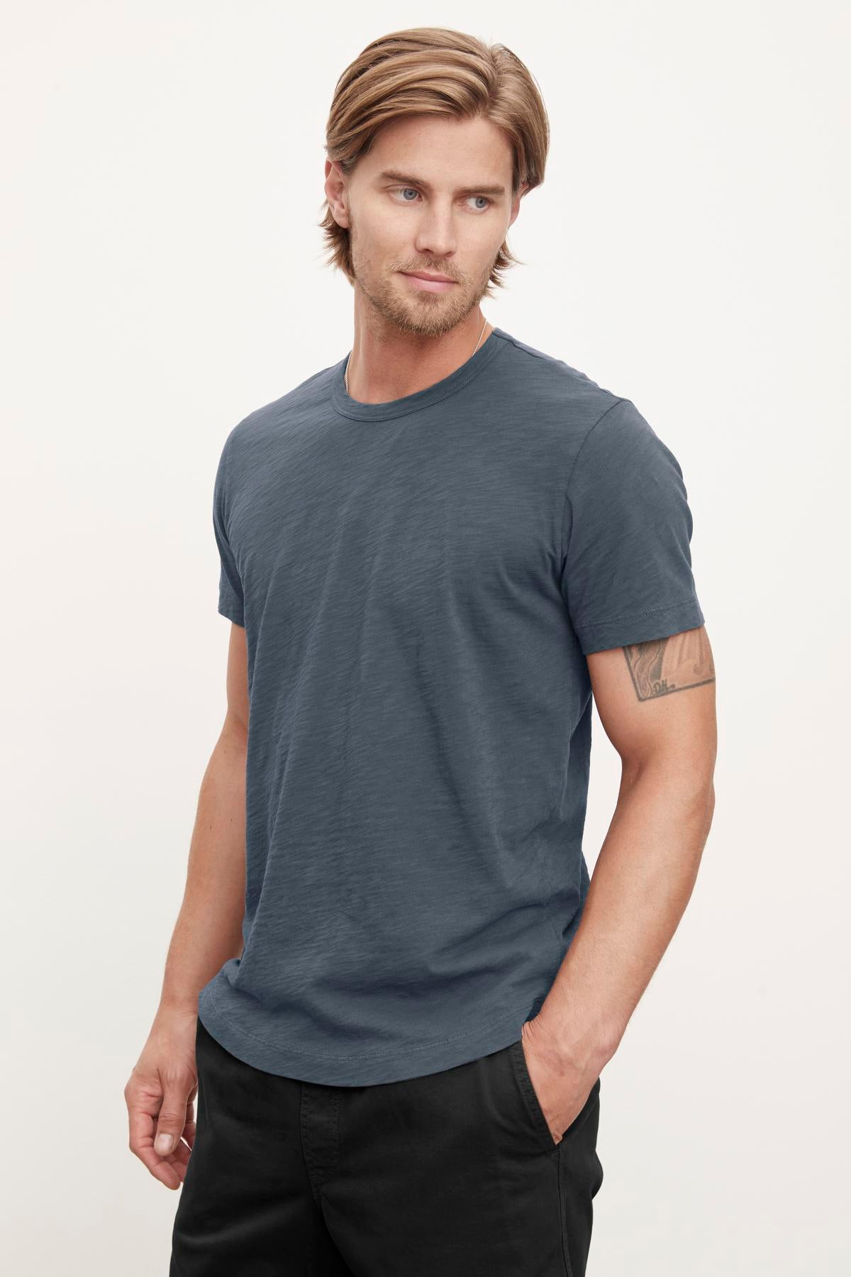 A man with medium-length hair wearing a blue Velvet by Graham & Spencer AMARO TEE and black pants stands with his left hand in his pocket, looking to his right, exuding a relaxed stylish feel.-37386214408385