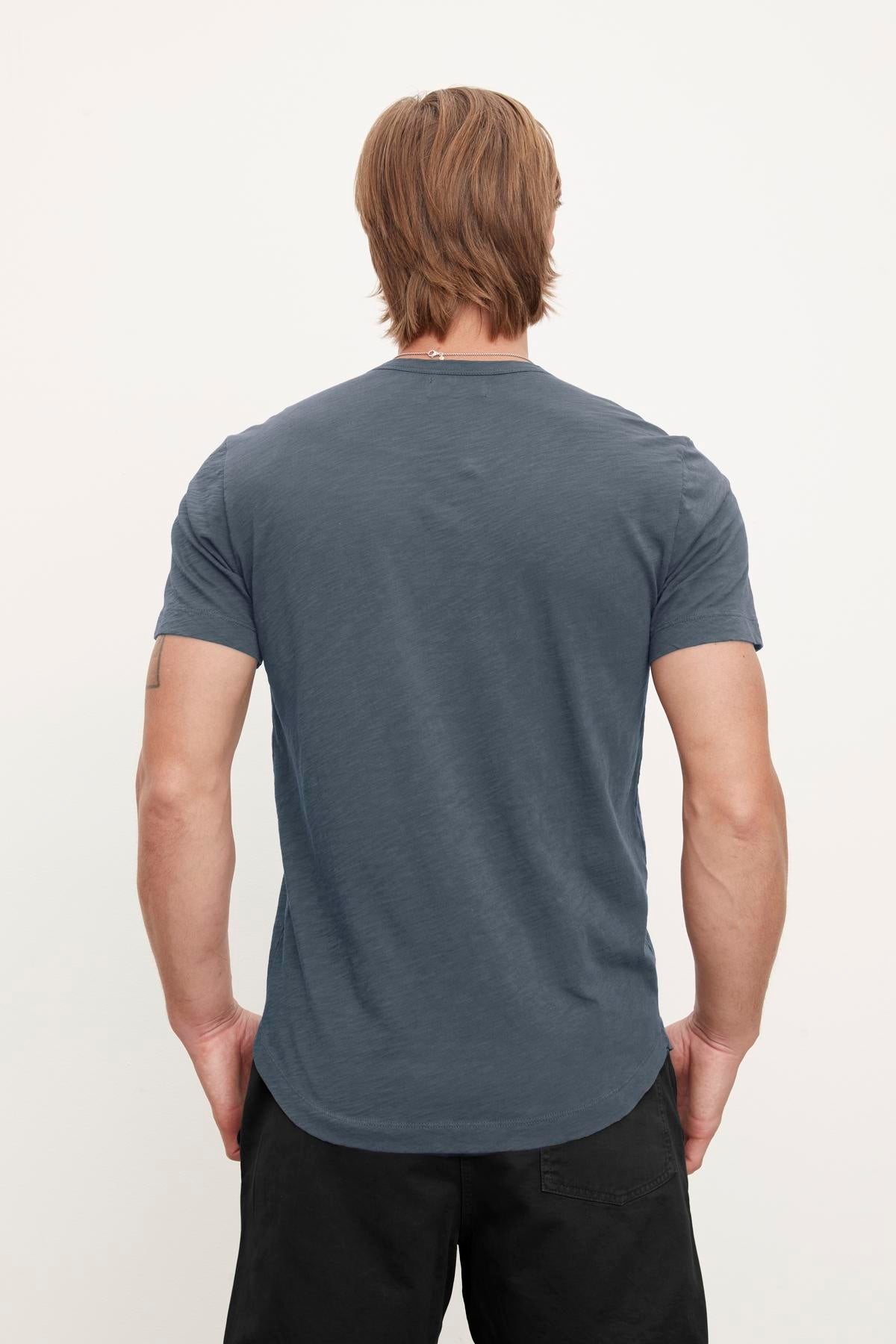 A person with shoulder-length hair is standing with their back to the camera, wearing a plain dark blue Velvet by Graham & Spencer AMARO TEE and black pants, exuding a relaxed stylish feel.-37386214441153
