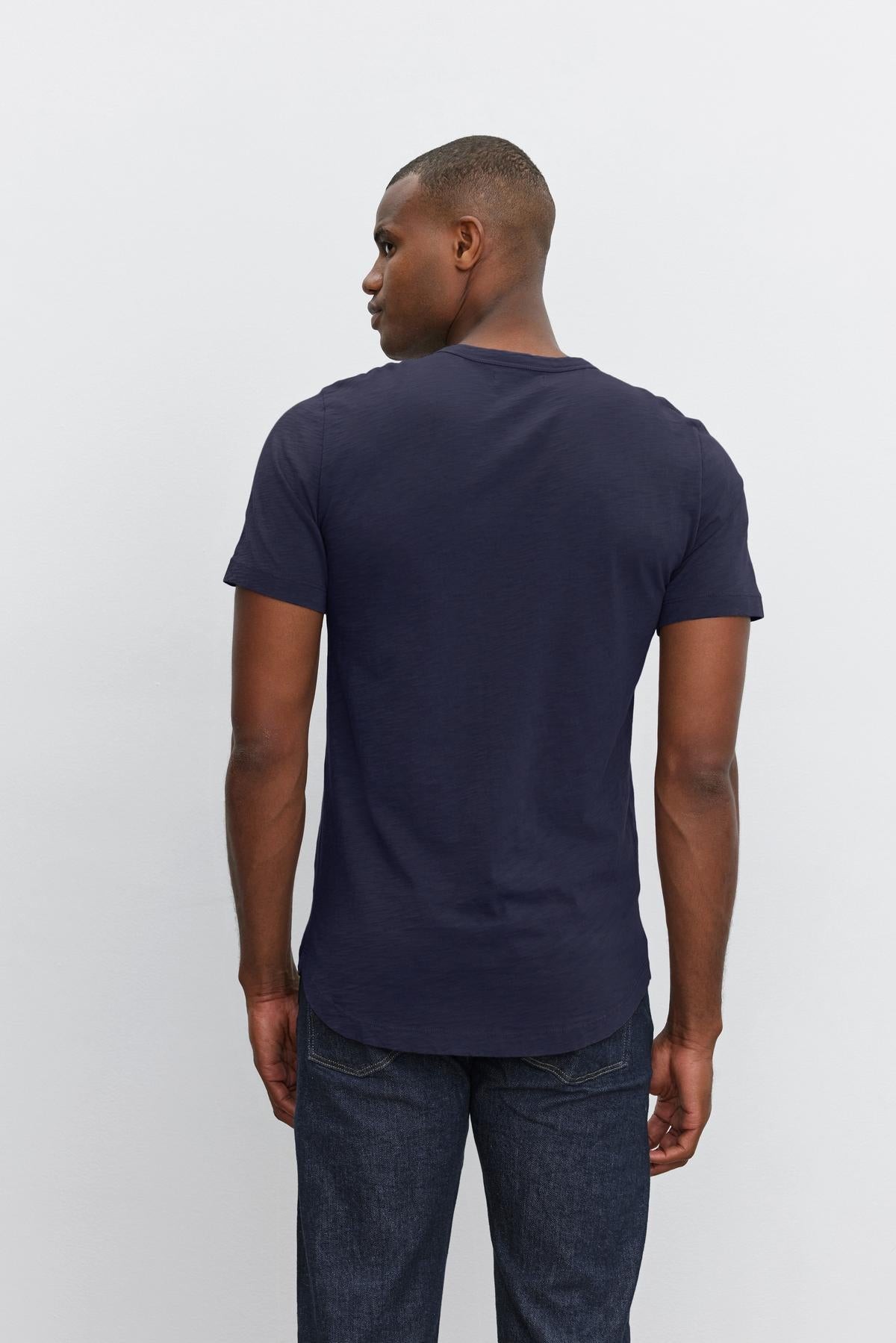 Man wearing a dark blue Velvet by Graham & Spencer crew neck AMARO TEE with heathered texture and denim jeans standing against a white background, viewed from behind.-36273921949889