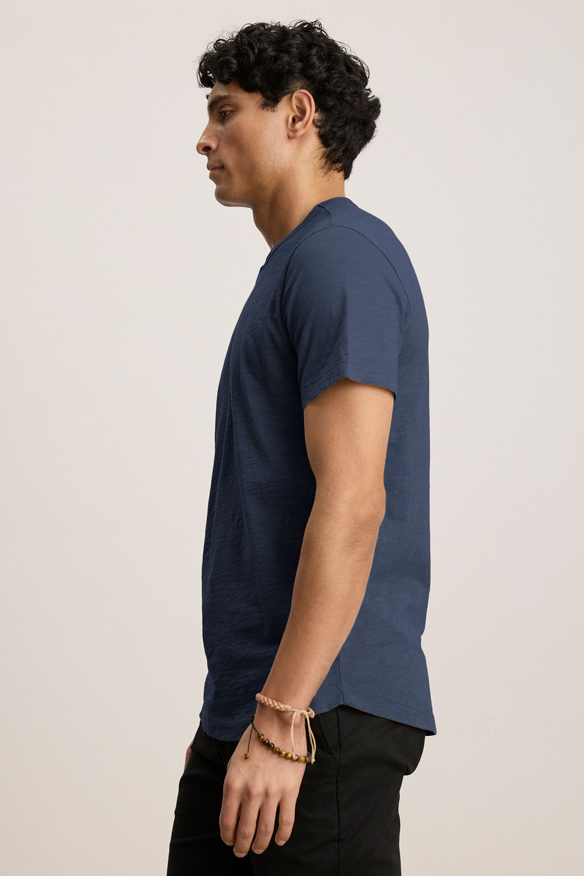   A person with dark hair, in a side profile view, wears a navy blue AMARO TEE by Velvet by Graham & Spencer featuring a classic crew neck and black pants against a plain light background. 