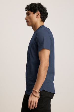 A person with dark hair, in a side profile view, wears a navy blue AMARO TEE by Velvet by Graham & Spencer featuring a classic crew neck and black pants against a plain light background.