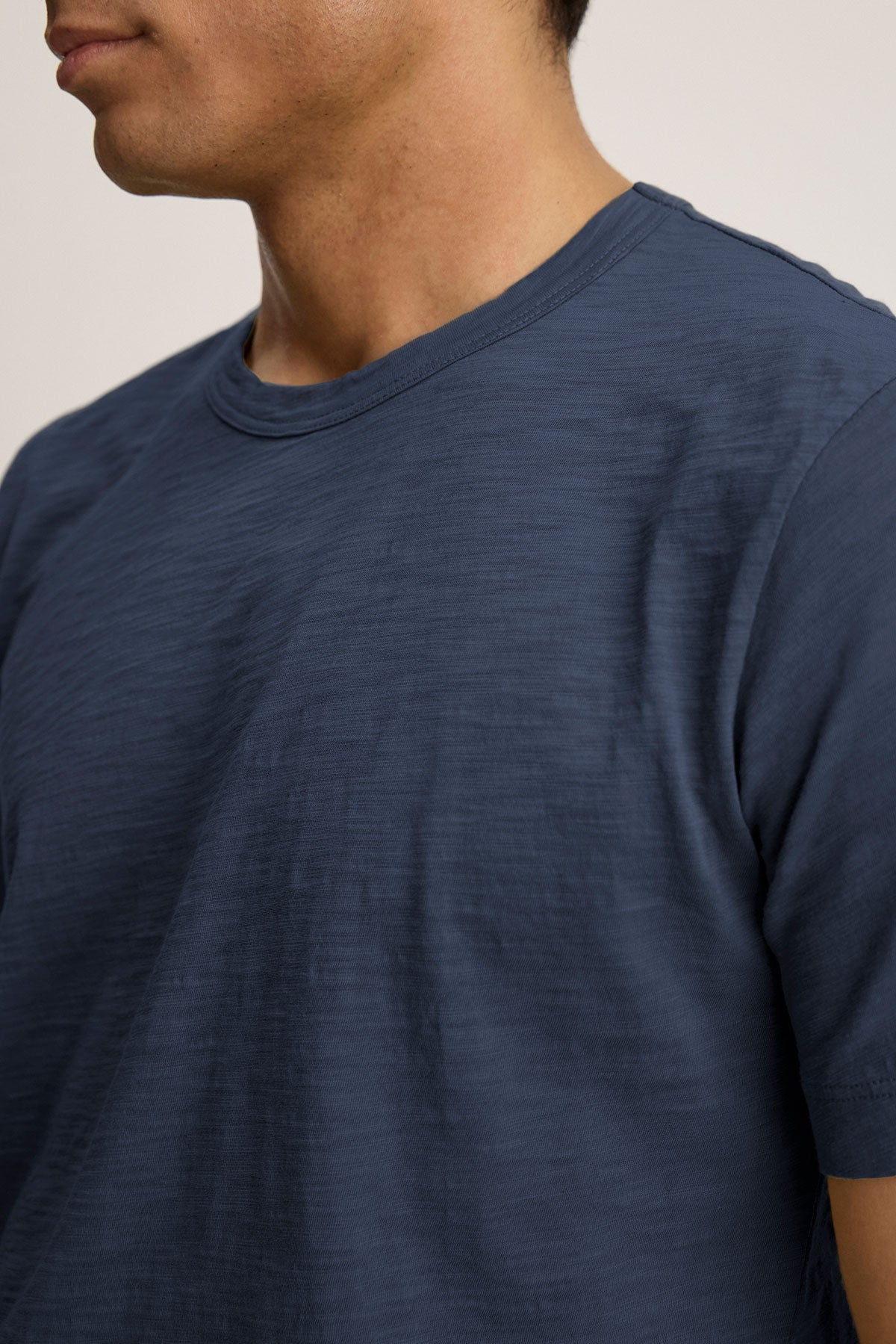 A person dons the AMARO TEE by Velvet by Graham & Spencer, a blue crew neck t-shirt that highlights style from shoulders to chest. The partly turned face emphasizes the tee's versatility as a layering piece crafted from soft Peruvian cotton.-38877608935617