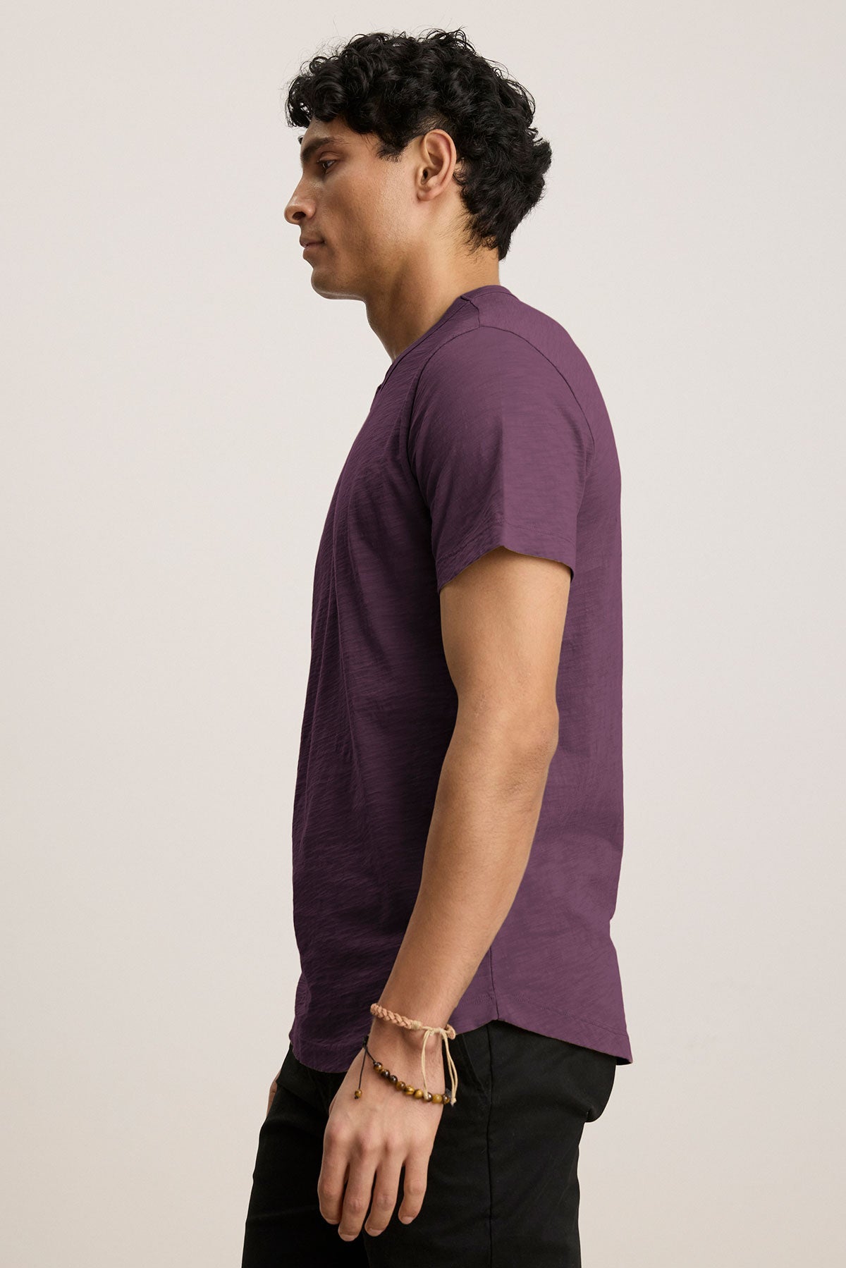   A person with curly hair stands in profile against a plain background, clad in a Velvet by Graham & Spencer AMARO TEE in purple, featuring a classic crew neckline. The black pants and bracelet complete the look, with the lightweight Peruvian cotton tee perfect for layering. 