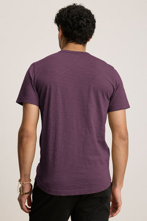 A person in a purple AMARO TEE by Velvet by Graham & Spencer, featuring a classic crew neckline, stands with their back to the camera. The lightweight Peruvian cotton makes it perfect for layering on any occasion. They are also wearing black pants.