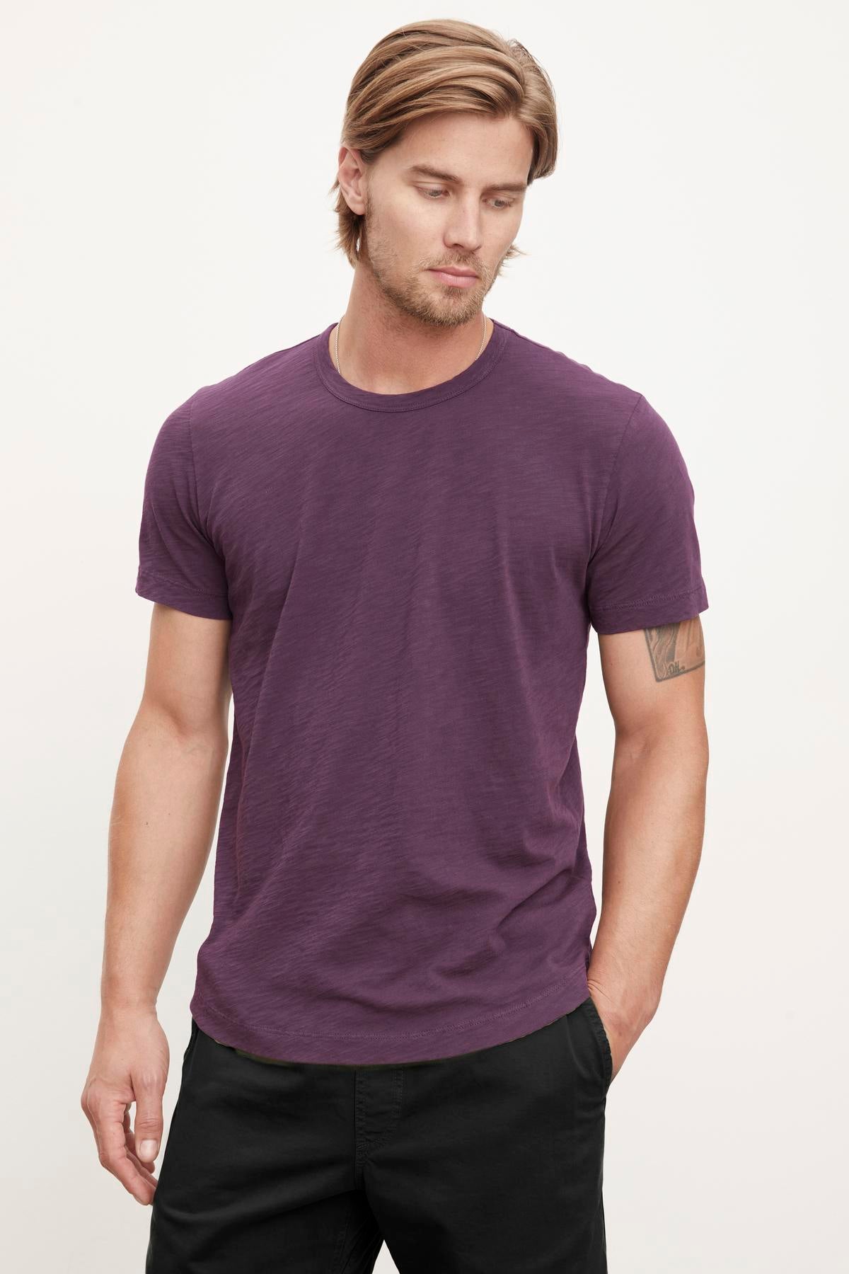 A person with light brown hair is wearing the AMARO TEE by Velvet by Graham & Spencer against a plain background, looking slightly downward. This purple t-shirt, featuring a classic crew neckline and crafted from lightweight Peruvian cotton, makes an excellent layering piece for any season.-38466905178305