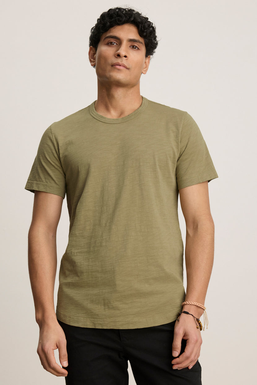 A person stands facing the camera against a plain light background, wearing the AMARO TEE by Velvet by Graham & Spencer—an olive green Peruvian cotton short-sleeve crew neck t-shirt—with black pants.
