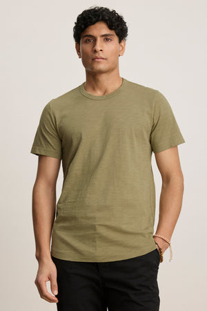A man stands facing forward in the AMARO TEE by Velvet by Graham & Spencer, an olive green Peruvian cotton t-shirt with a classic crew neckline, paired with black pants against a neutral background.