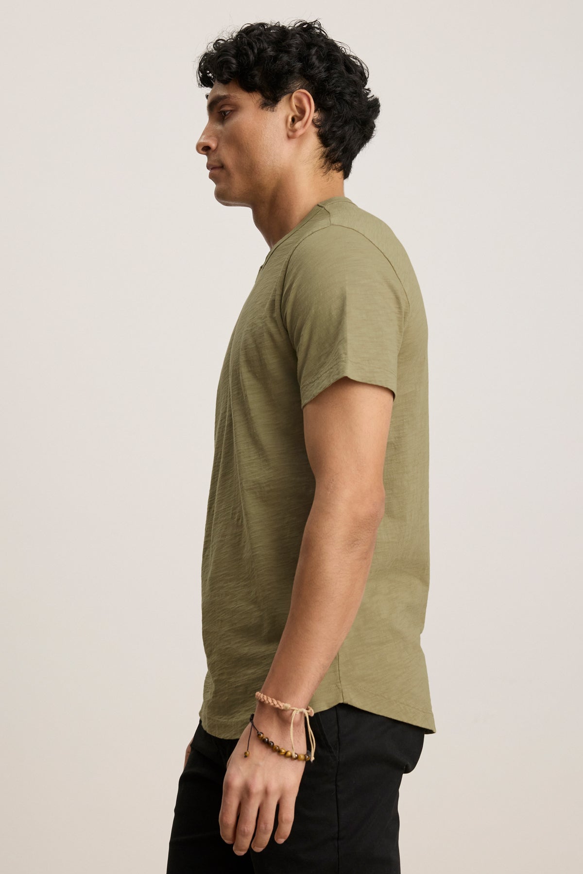 A man stands in profile wearing the AMARO TEE by Velvet by Graham & Spencer—an olive green Peruvian cotton short-sleeve crewneck—paired with black pants and a beaded bracelet, against a plain background.-39680467632321