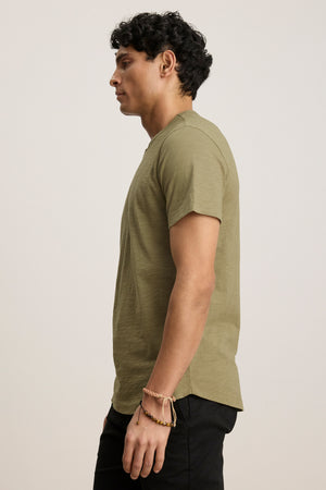 A man stands in profile wearing the AMARO TEE by Velvet by Graham & Spencer—an olive green Peruvian cotton short-sleeve crewneck—paired with black pants and a beaded bracelet, against a plain background.
