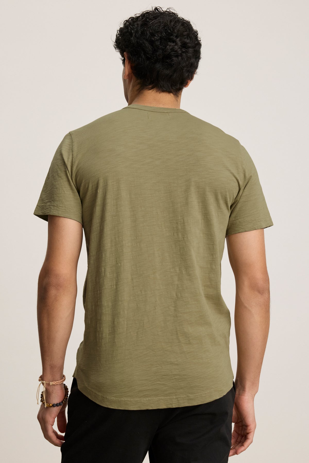 A person with short curly hair is shown from the back, wearing the AMARO TEE in olive green by Velvet by Graham & Spencer and black pants against a plain background.-39680467599553