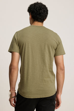 A person with short curly hair is shown from the back, wearing the AMARO TEE in olive green by Velvet by Graham & Spencer and black pants against a plain background.