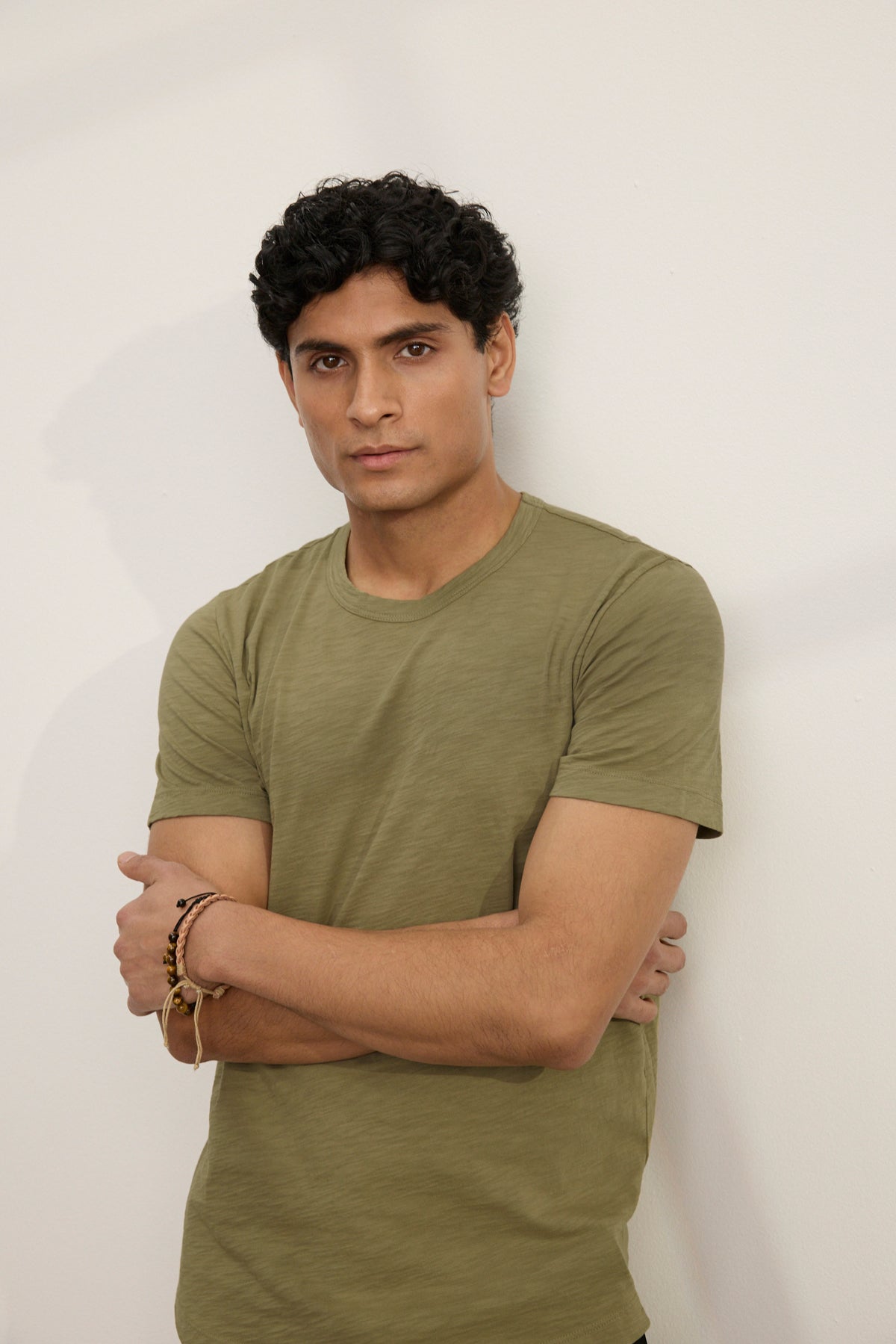 A man with short curly hair stands against a plain light-colored wall, arms crossed, wearing the AMARO TEE by Velvet by Graham & Spencer—an olive green Peruvian cotton crew neck t-shirt.-39680467534017