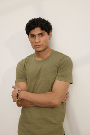 A man with short curly hair stands against a plain light-colored wall, arms crossed, wearing the AMARO TEE by Velvet by Graham & Spencer—an olive green Peruvian cotton crew neck t-shirt.