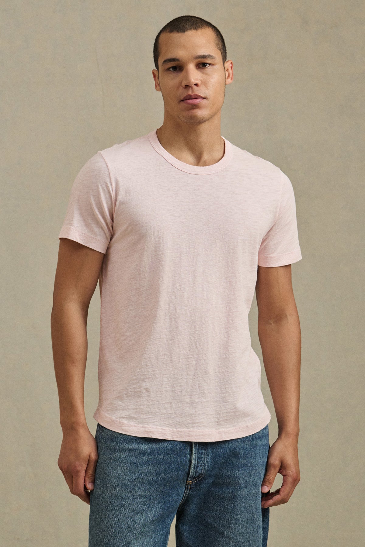 A man stands against a neutral background, wearing the Velvet by Graham & Spencer AMARO TEE in light pink Peruvian cotton with a crew neckline and blue jeans—an ideal layering piece for any wardrobe.