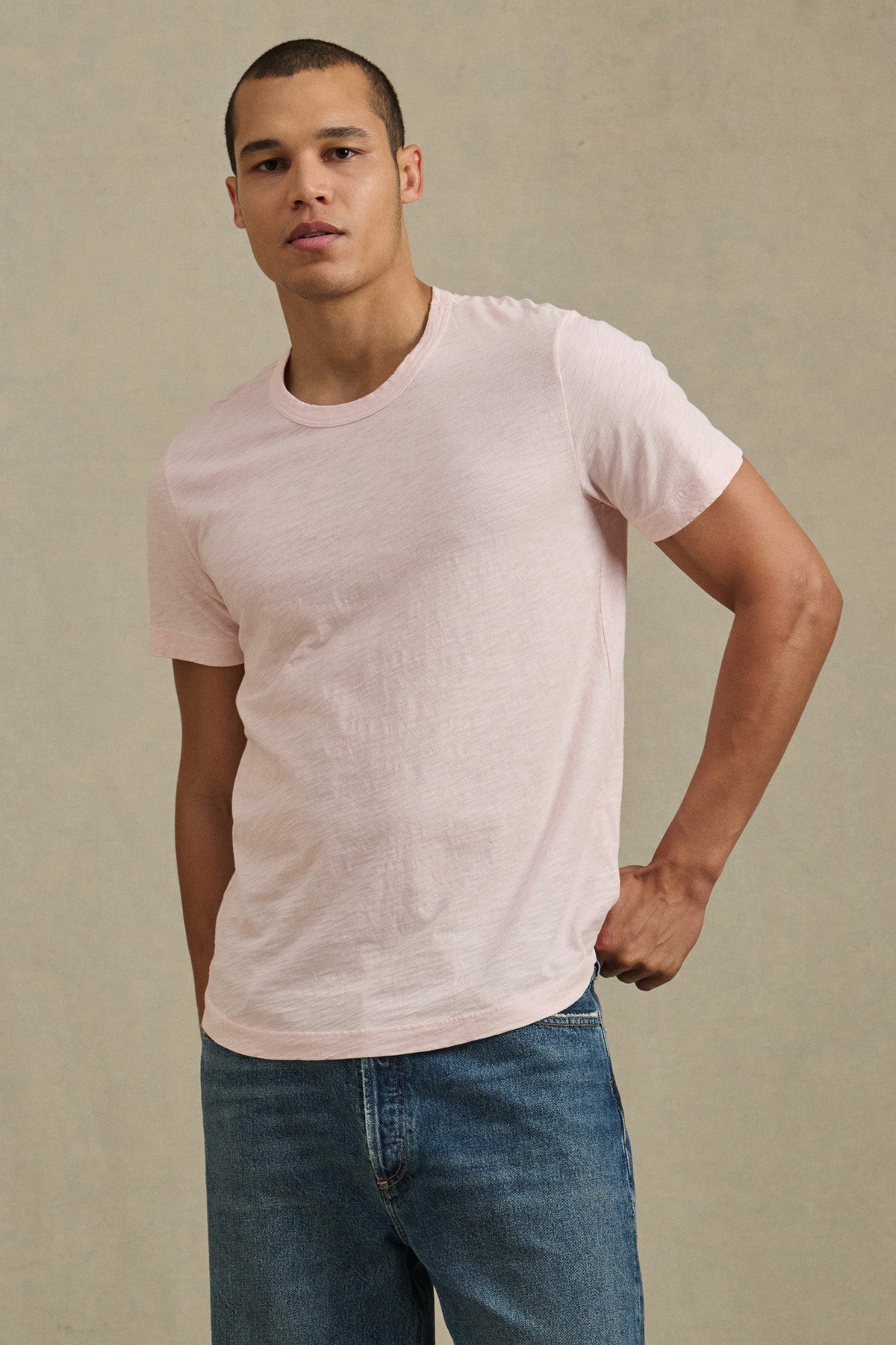 A man poses against a plain background, wearing the AMARO TEE by Velvet by Graham & Spencer—a light pink Peruvian cotton T-shirt with a classic crew neckline—and blue jeans, looking at the camera with one hand on his hip.-41008903684289