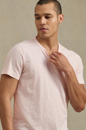 A man wearing the Velvet by Graham & Spencer AMARO TEE, a light pink short-sleeve crew neck, stands against a neutral background, looking down and to the side while touching his collar.