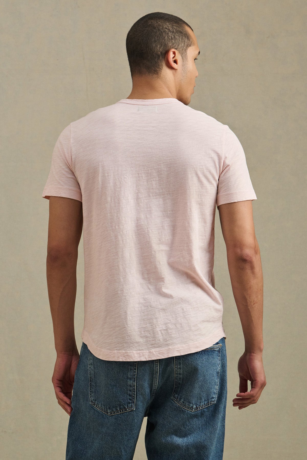 A man stands with his back to the camera, wearing the AMARO TEE by Velvet by Graham & Spencer—a light pink Peruvian cotton crewneck short-sleeve t-shirt—and blue jeans, set against a plain beige background.-41008903618753