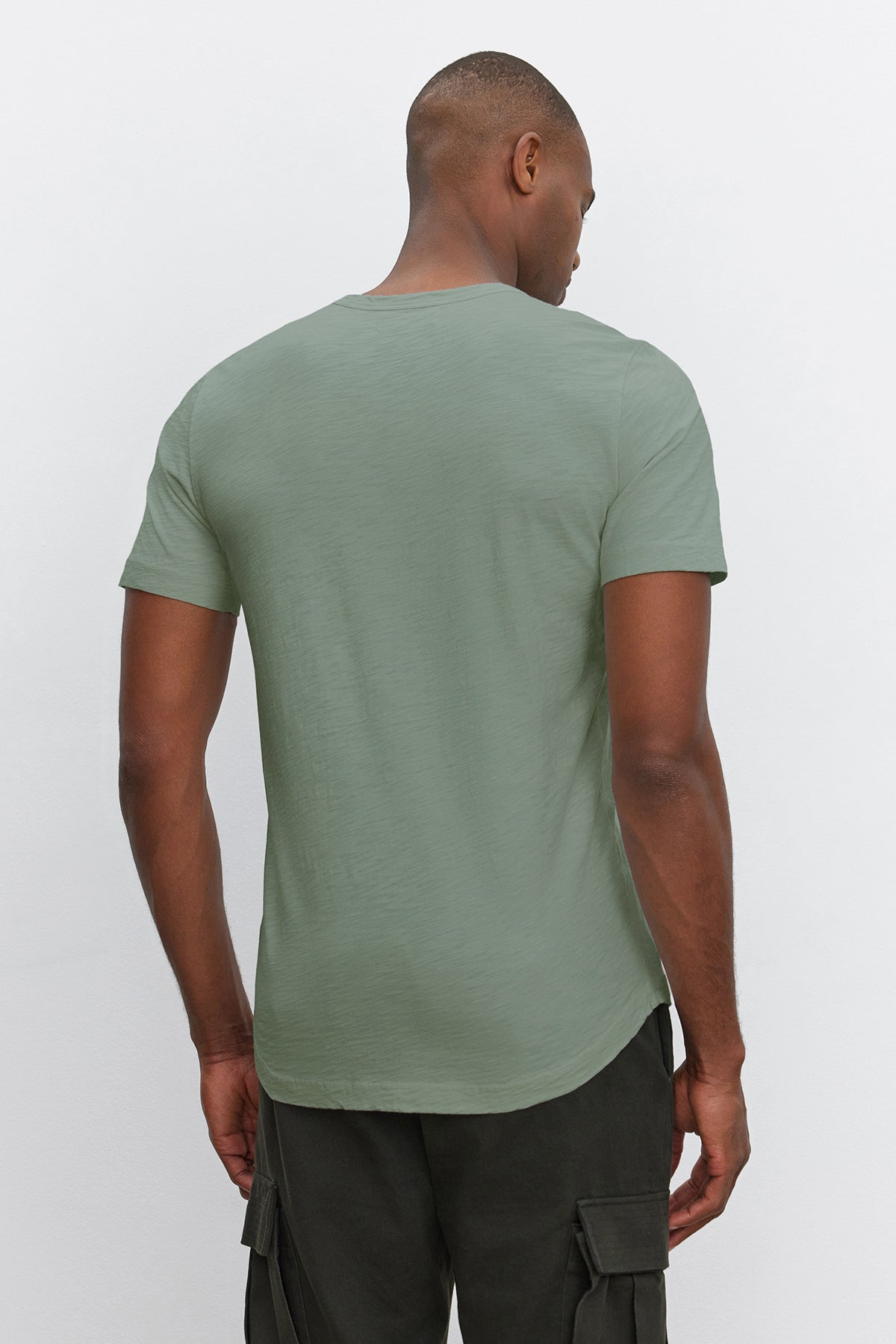 A man is standing with his back facing the camera, wearing a stylish light green Velvet by Graham & Spencer AMARO TEE and dark cargo pants.-36909384728769