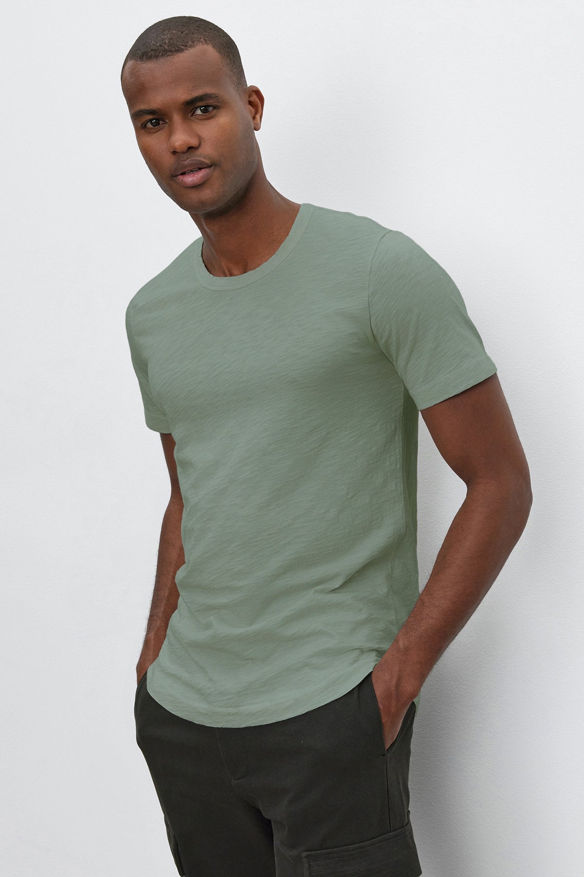A man wearing a stylish, medium-weight AMARO TEE by Velvet by Graham & Spencer in light green and dark pants stands against a white background, with hands in his pockets.-36909384761537