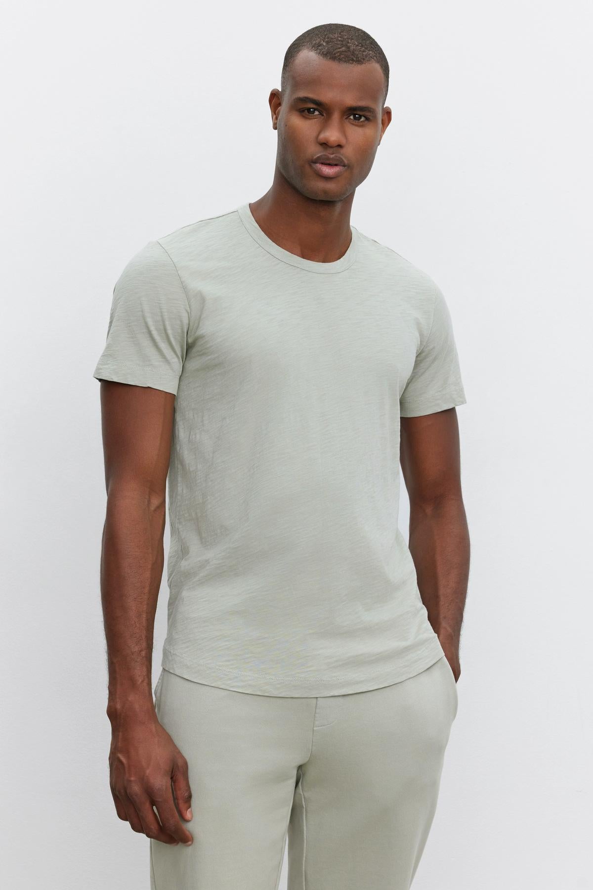 A person with a neutral expression stands against a plain white background, wearing a light grey AMARO TEE by Velvet by Graham & Spencer and matching grey pants, showcasing a versatile look with a relaxed and stylish feel.-37469132095681