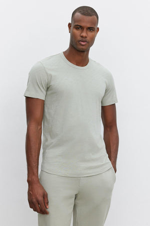 A person with a neutral expression stands against a plain white background, wearing a light grey AMARO TEE by Velvet by Graham & Spencer and matching grey pants, showcasing a versatile look with a relaxed and stylish feel.