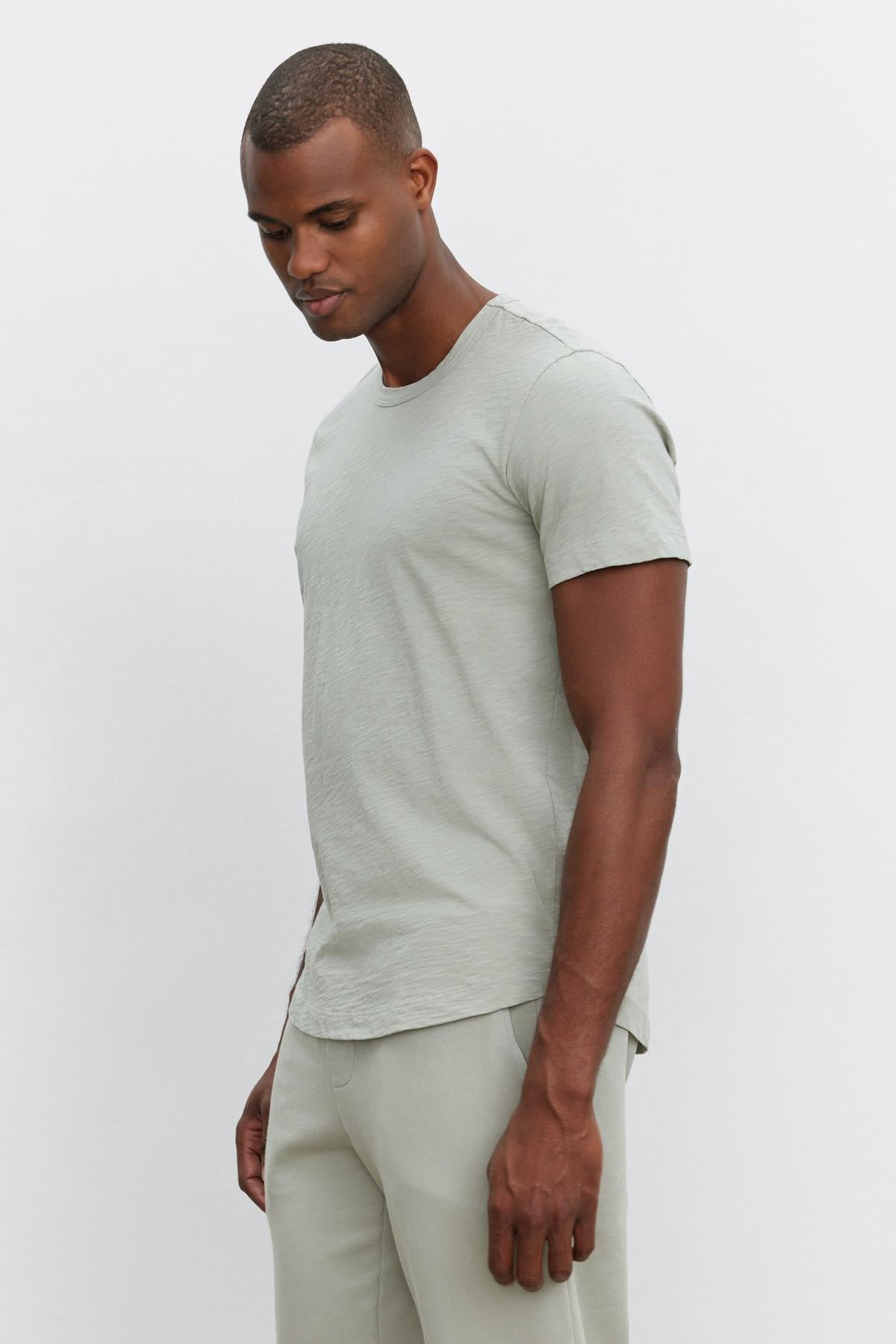 A man in a light grey Velvet by Graham & Spencer AMARO TEE and matching pants stands sideways against a plain white background, looking down, embodying a relaxed and stylish feel.-37469132128449