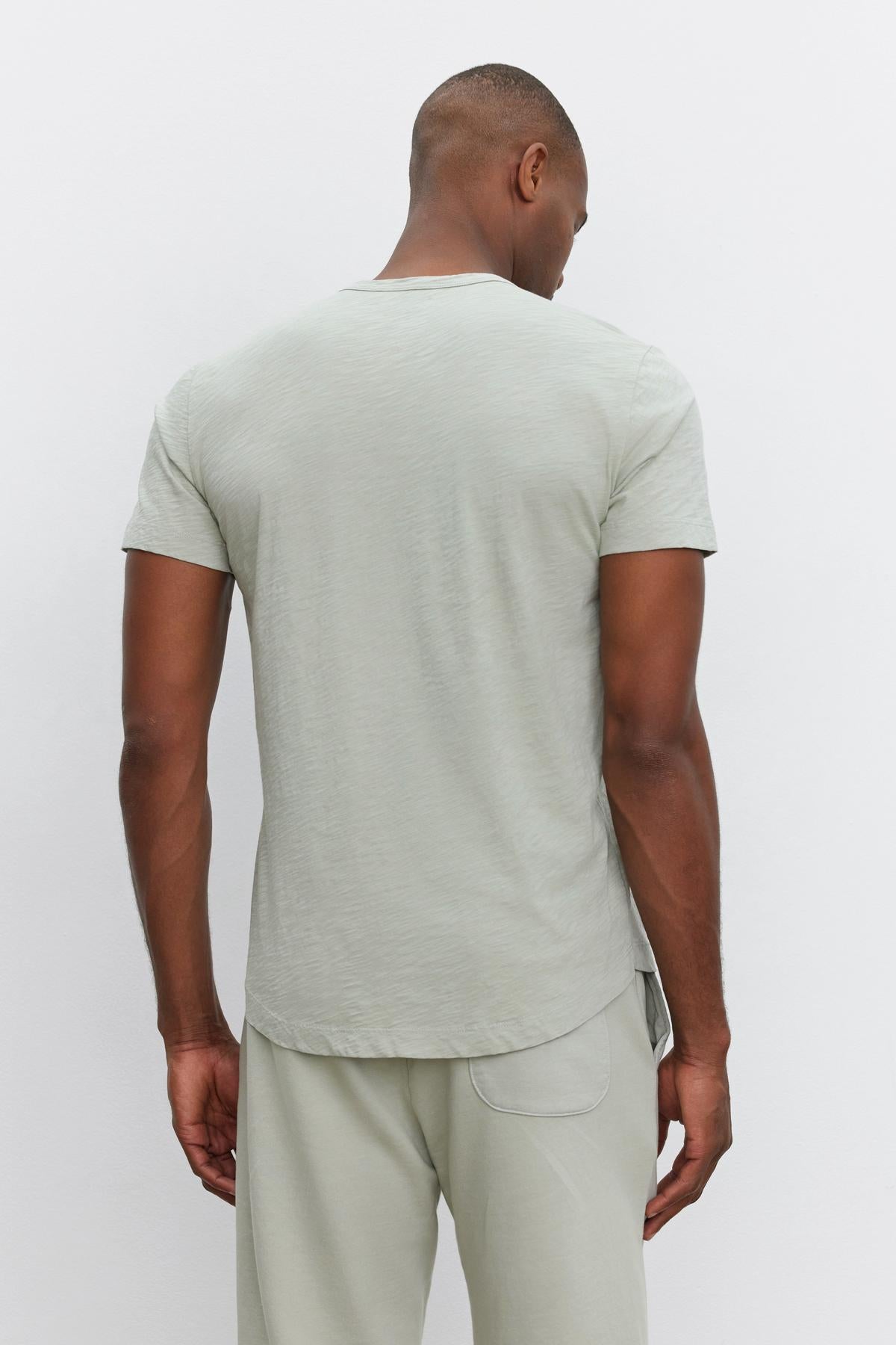 A person stands facing away, wearing a light gray short-sleeve AMARO TEE by Velvet by Graham & Spencer and matching pants, exuding a relaxed and stylish feel.-37469132161217