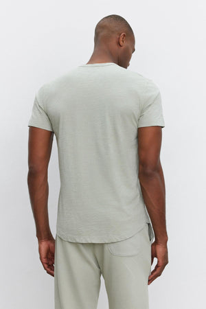 A person stands facing away, wearing a light gray short-sleeve AMARO TEE by Velvet by Graham & Spencer and matching pants, exuding a relaxed and stylish feel.