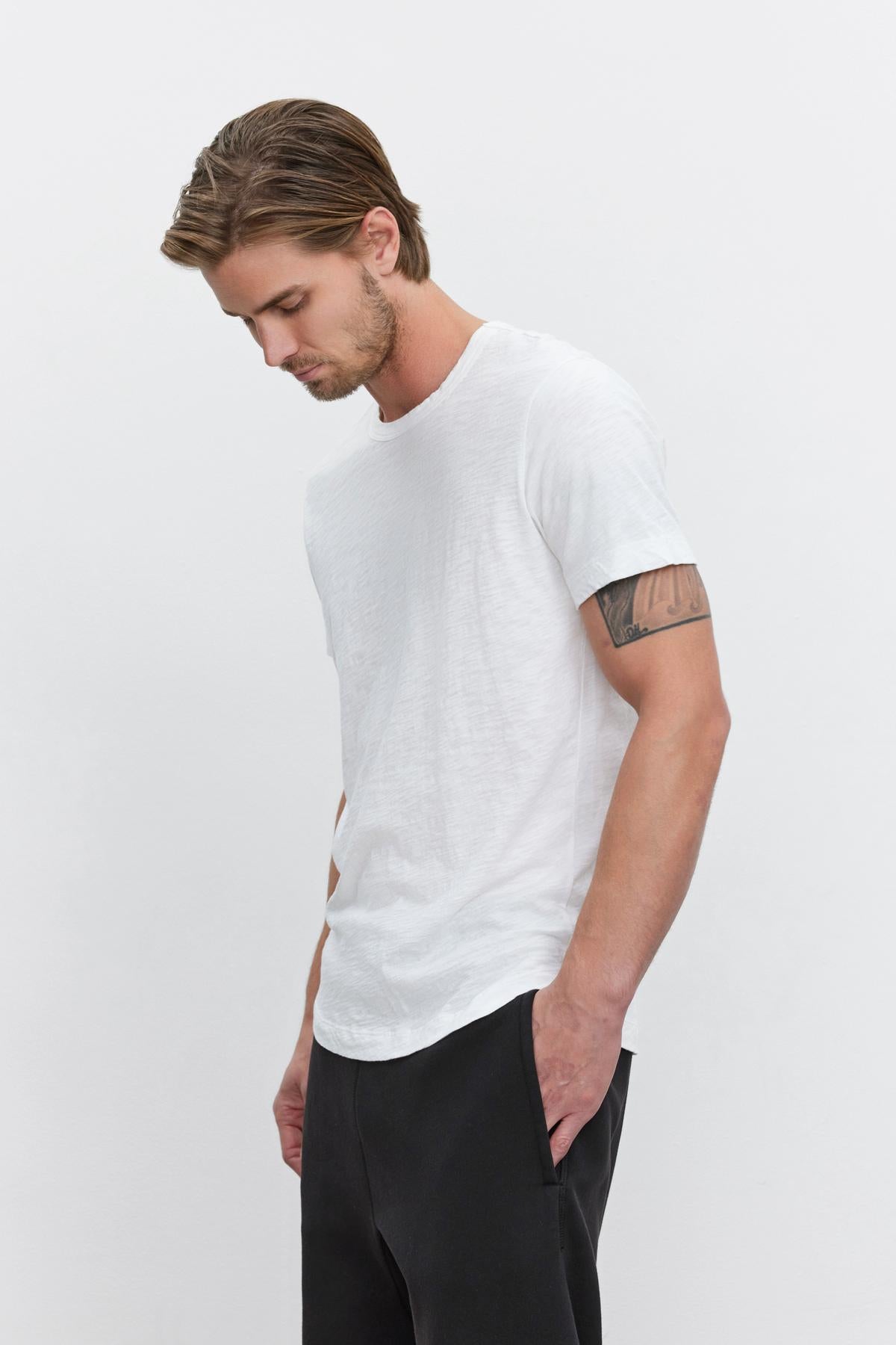 Man in a white crew neck Velvet by Graham & Spencer AMARO TEE with a heathered texture and black pants, looking downwards with a tattoo visible on his left arm.-35867457552577