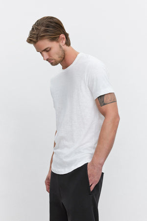 Man in a white crew neck Velvet by Graham & Spencer AMARO TEE with a heathered texture and black pants, looking downwards with a tattoo visible on his left arm.