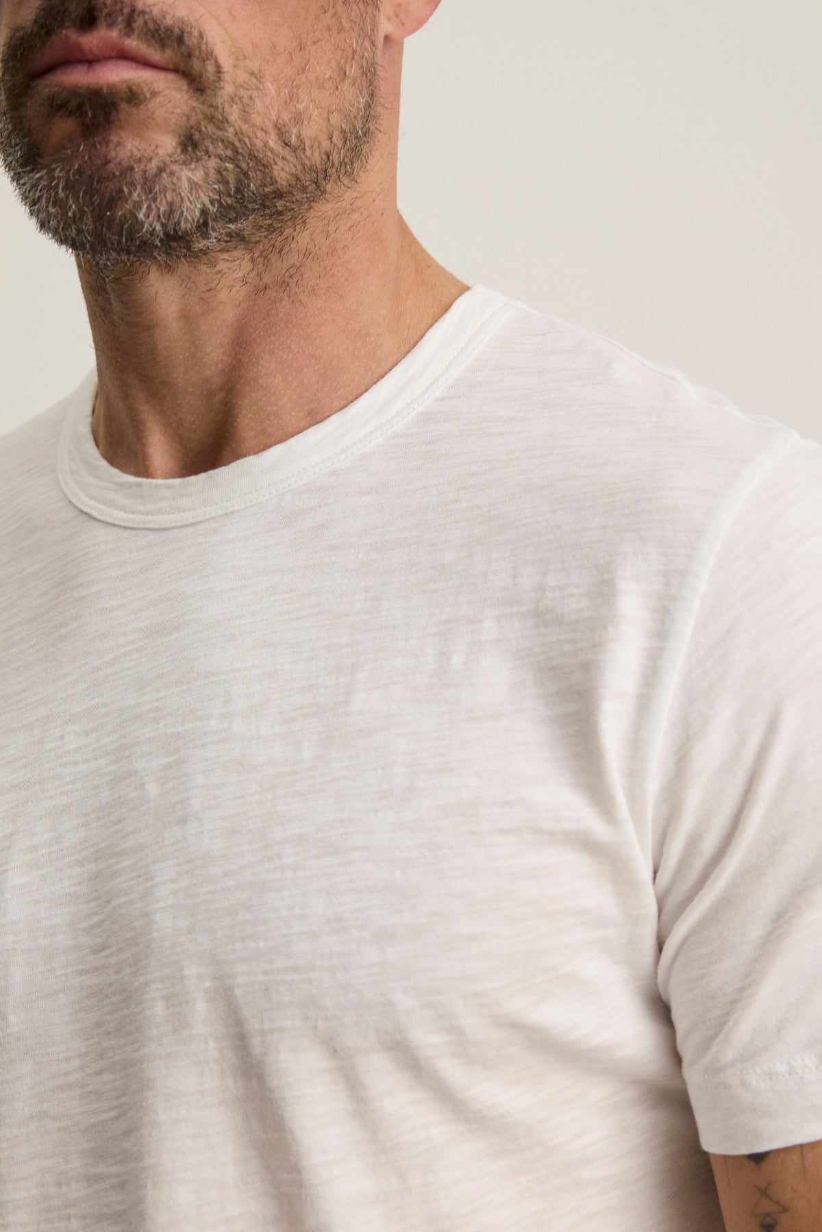   A bearded person wears the AMARO TEE by Velvet by Graham & Spencer, a plain white T-shirt made from soft Peruvian cotton with a classic crew neckline—perfect for versatile layering. 