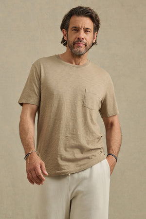 A man with short brown hair and a beard wears the Velvet by Graham & Spencer BRETT TEE, a beige T-shirt with a chest pocket, paired with off-white pants, standing against a plain beige background.