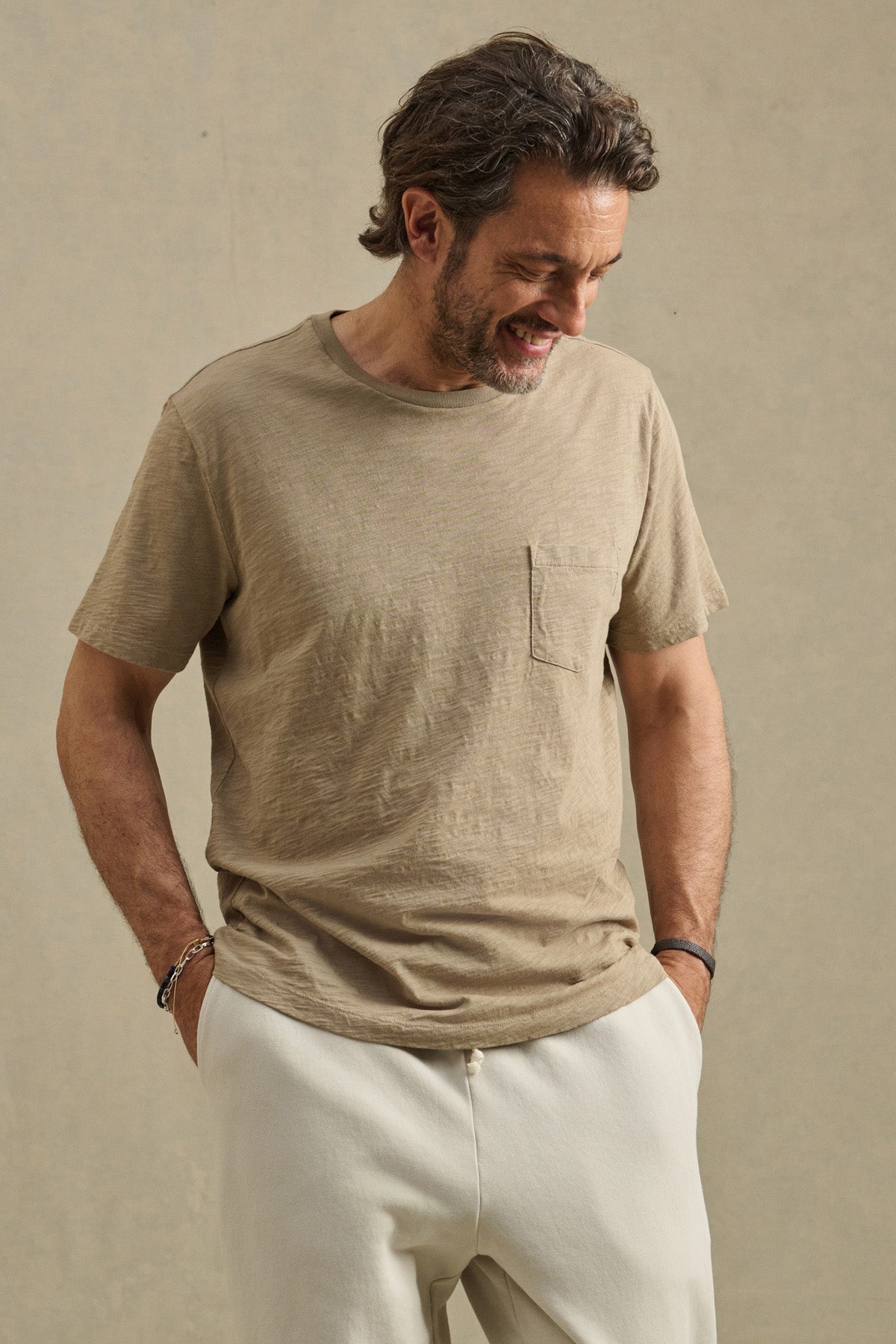 Man wearing the BRETT TEE by Velvet by Graham & Spencer—a light brown short-sleeve t-shirt with a chest pocket—paired with cream-colored pants, stands against a beige background, looking down and smiling.