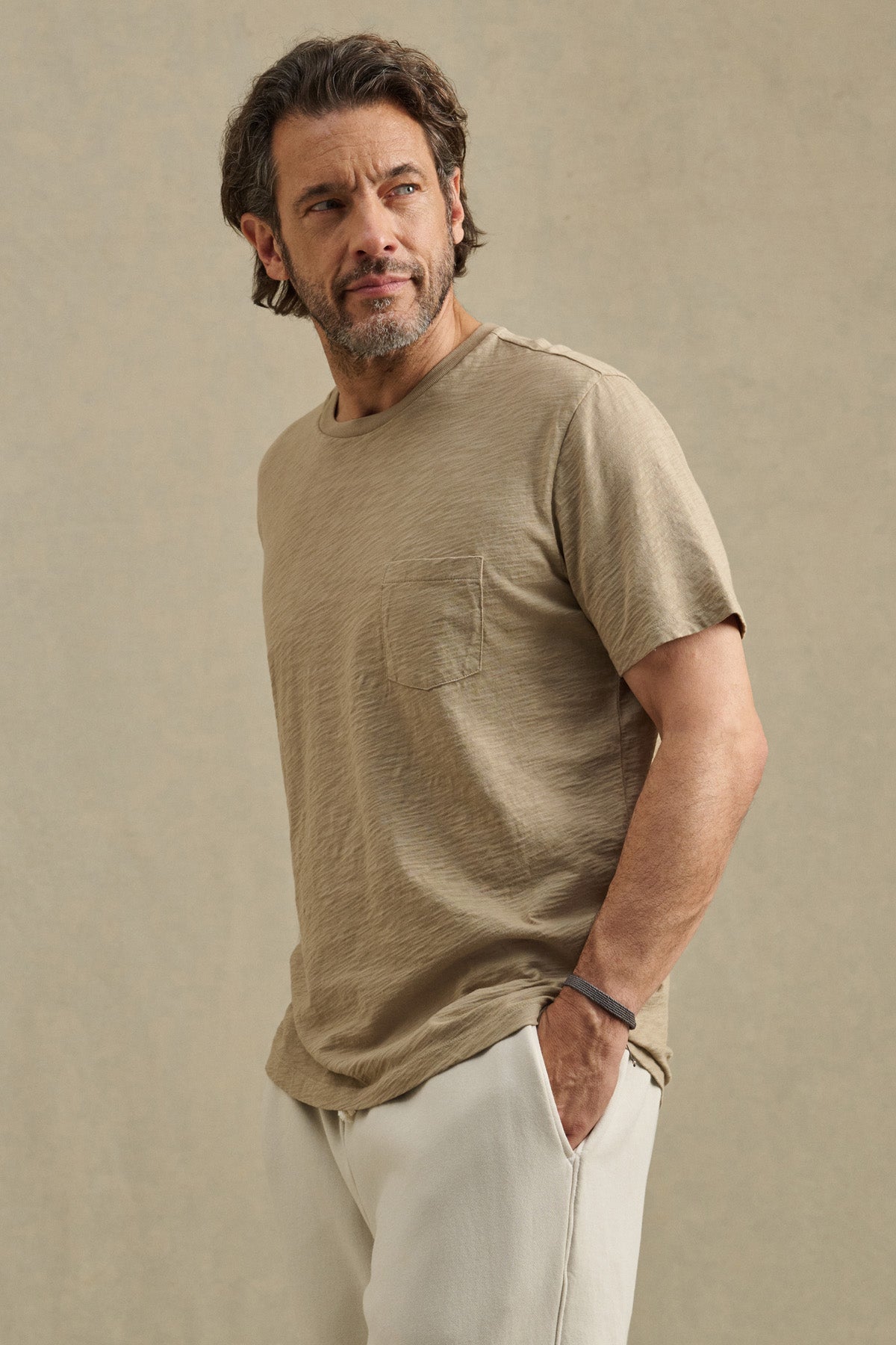 A man with medium-length hair and a beard wears the BRETT TEE by Velvet by Graham & Spencer in beige, paired with light pants, standing against a plain beige background with one hand in his pocket.