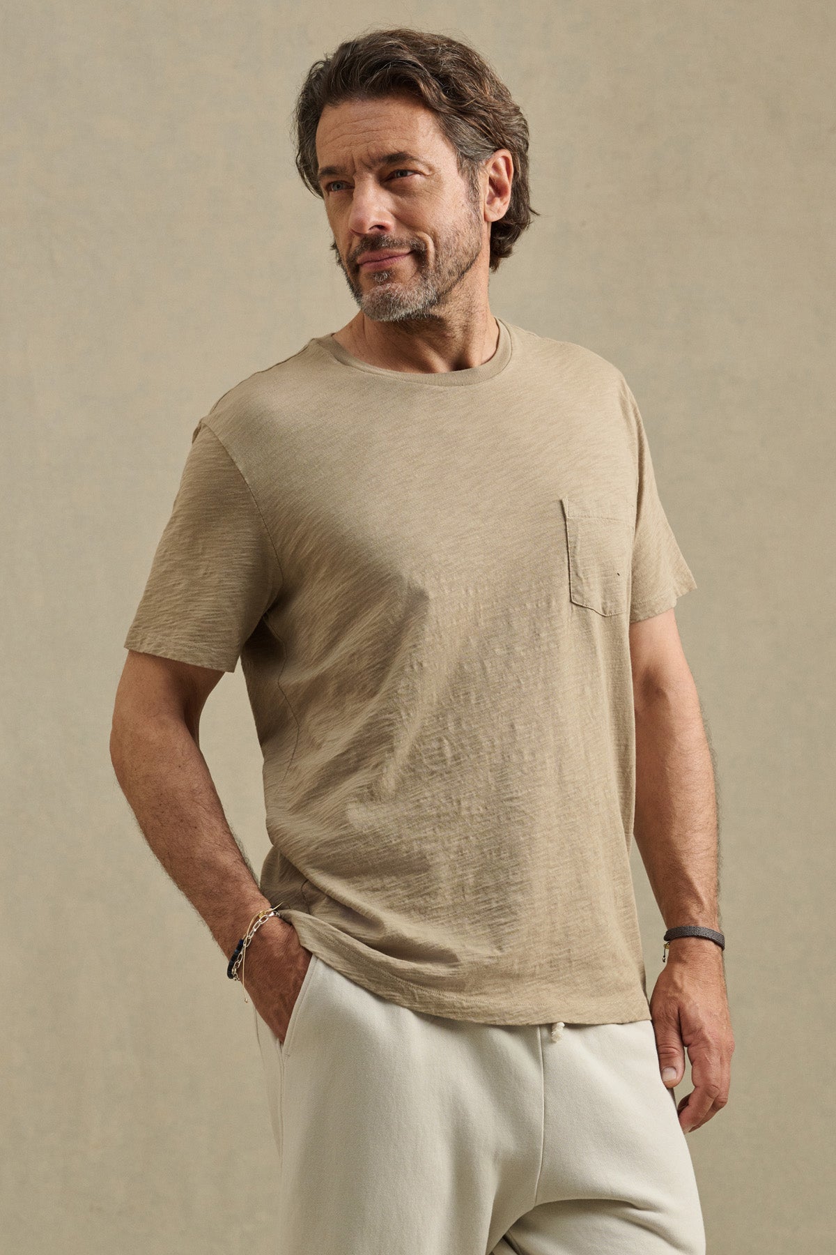 A man wears the BRETT TEE by Velvet by Graham & Spencer, a beige short-sleeve t-shirt with a chest pocket, paired with light-colored pants, standing against a plain neutral background.-41374364598465