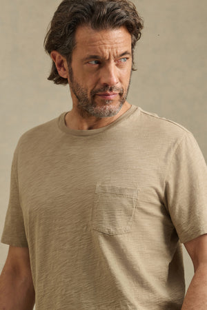 A man with medium-length brown hair and a beard wears the BRETT TEE by Velvet by Graham & Spencer—a beige t-shirt with a chest pocket—as he looks left against a neutral background.