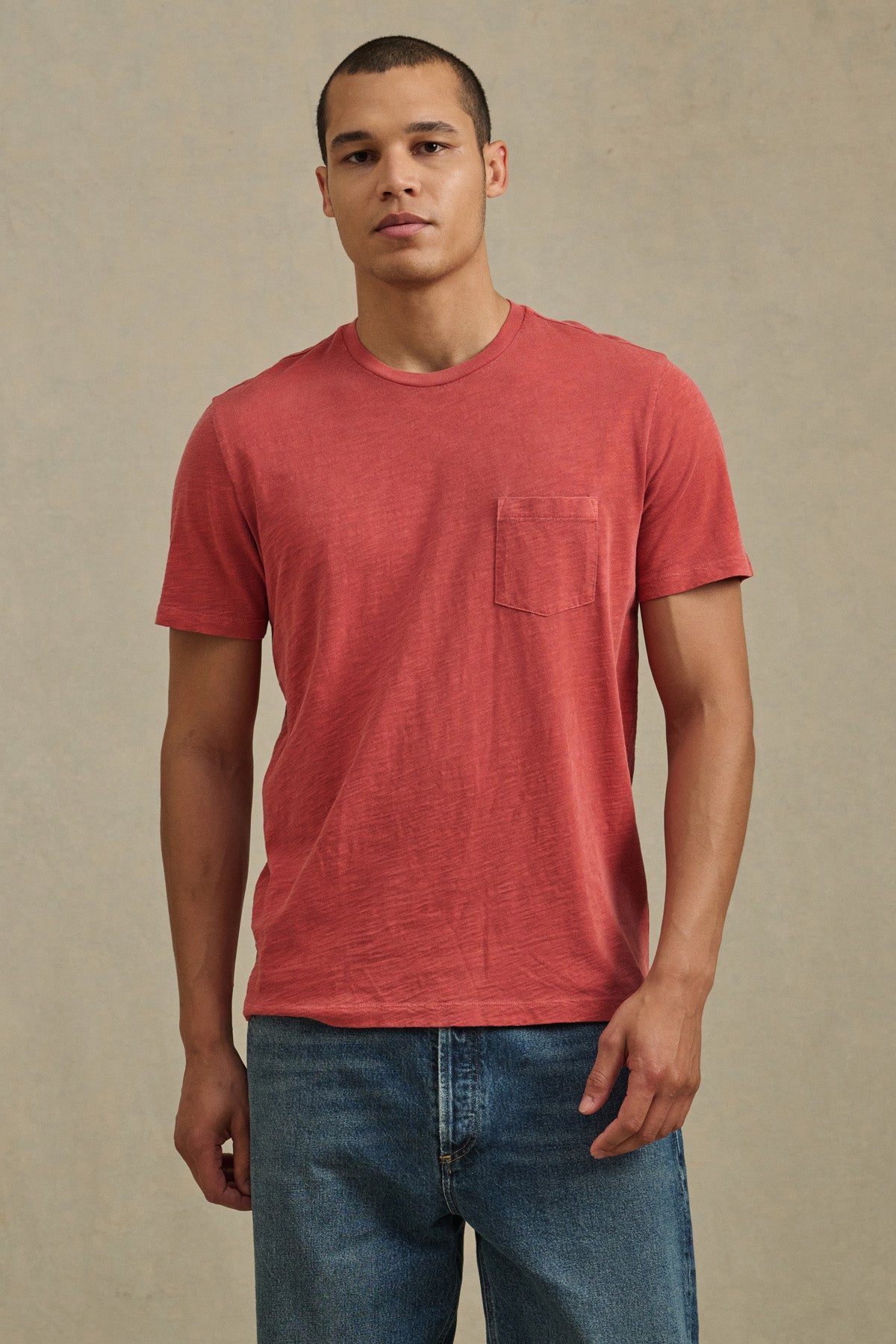 A man faces forward wearing the Velvet by Graham & Spencer BRETT TEE, a red short-sleeve T-shirt with a chest pocket, paired with blue jeans against a neutral background.-41374364696769