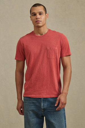 A man faces forward wearing the Velvet by Graham & Spencer BRETT TEE, a red short-sleeve T-shirt with a chest pocket, paired with blue jeans against a neutral background.