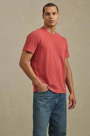 A man wears the BRETT TEE by Velvet by Graham & Spencer—a red short-sleeve t-shirt with a chest pocket—paired with blue jeans, standing against a plain beige background.