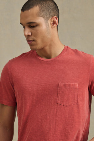A person with short hair looks down against a plain background, wearing the BRETT TEE by Velvet by Graham & Spencer—a faded red t-shirt featuring a chest pocket.