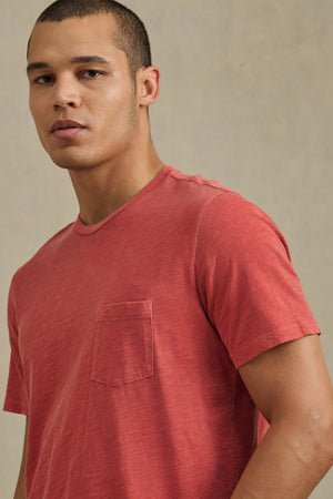 A man with short hair wears the BRETT TEE by Velvet by Graham & Spencer—a faded red t-shirt with a chest pocket—while standing against a neutral background, looking slightly past the camera.