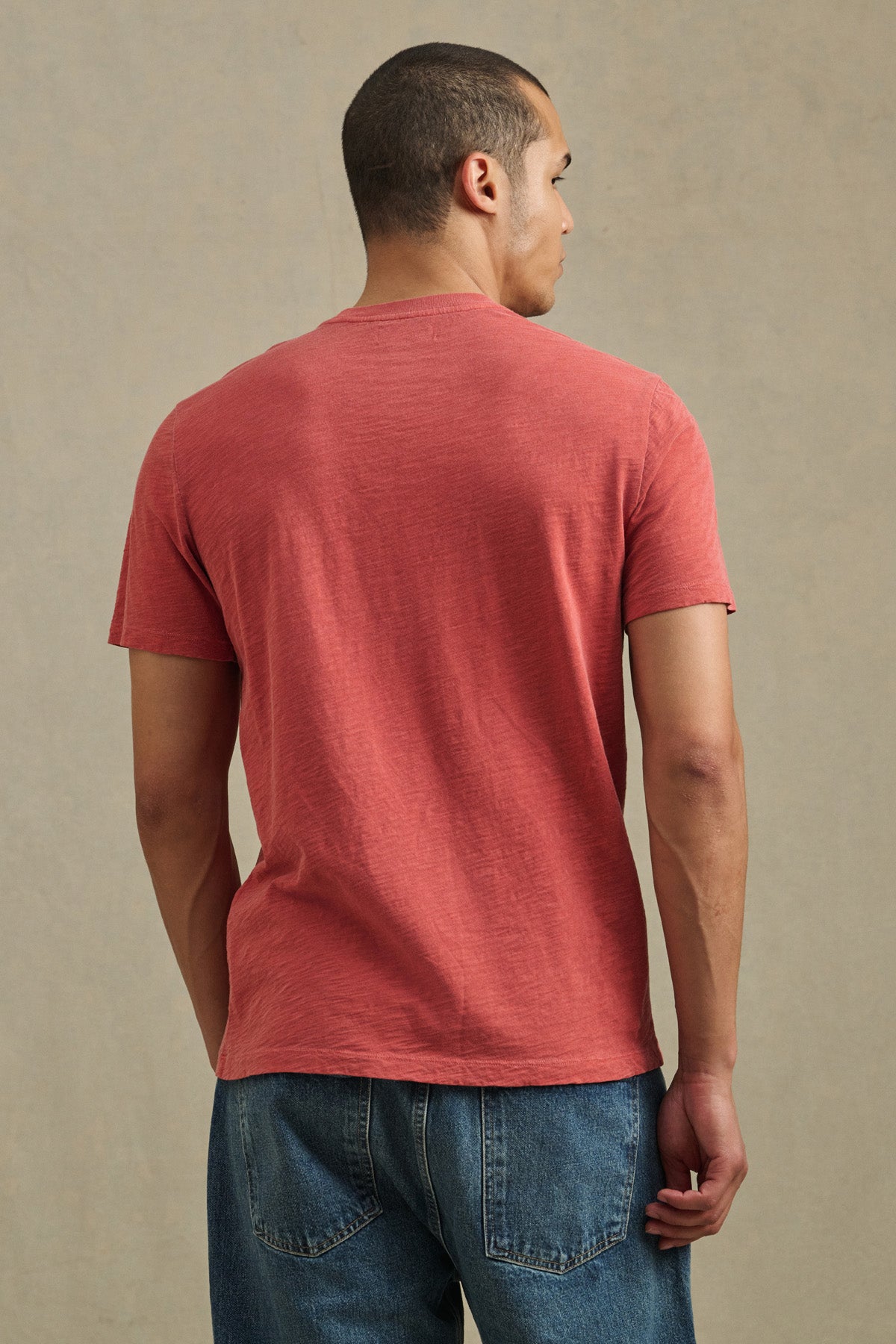 A man stands with his back to the camera, wearing the Velvet by Graham & Spencer BRETT TEE in red and blue jeans against a neutral background.