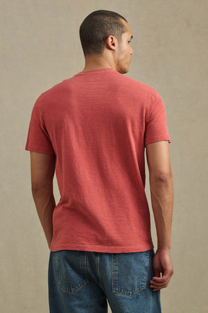 A man stands with his back to the camera, wearing the Velvet by Graham & Spencer BRETT TEE in red and blue jeans against a neutral background.