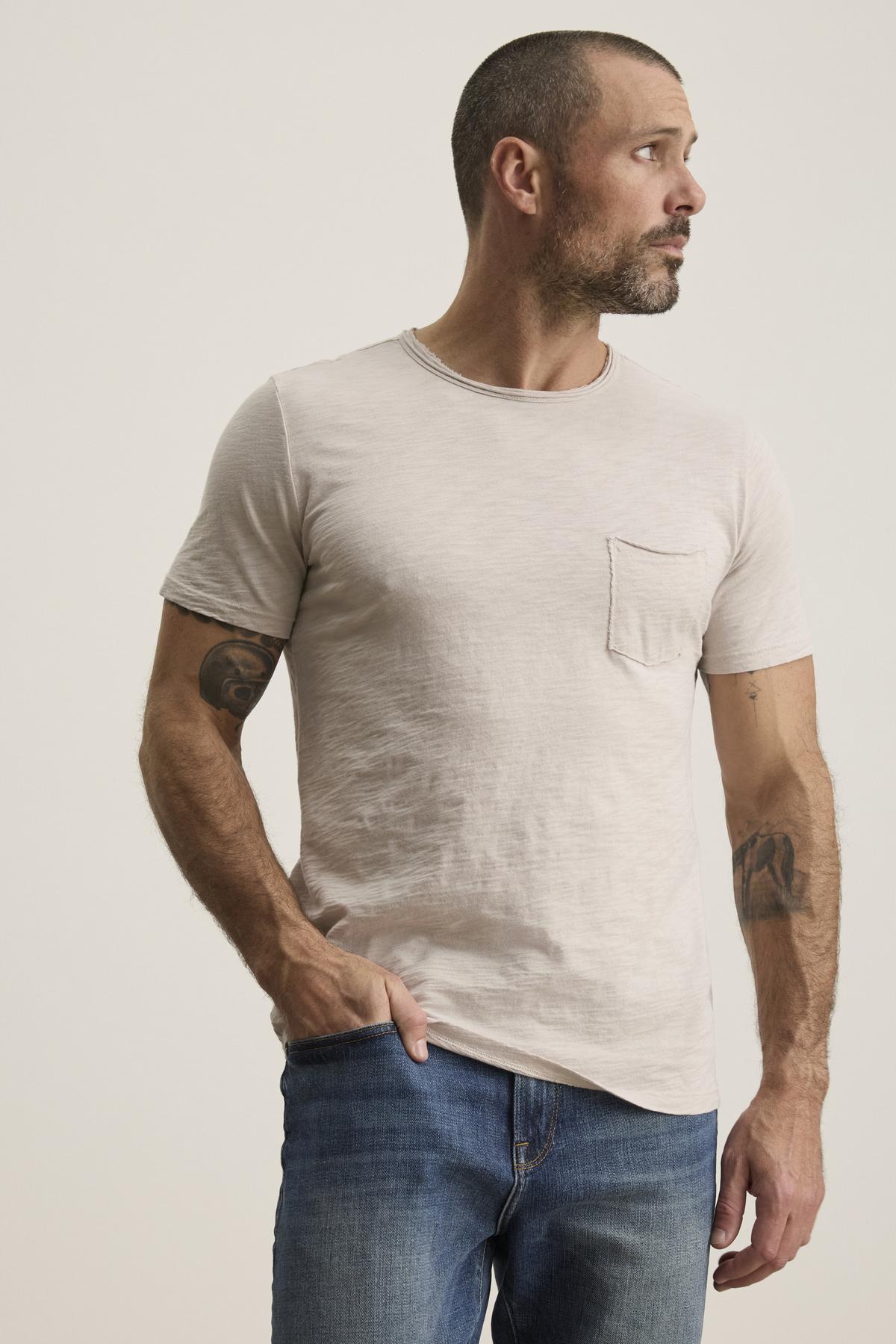   A man with short hair and a beard is wearing the CHAD TEE from Velvet by Graham & Spencer, made of soft Peruvian cotton in light beige, paired with blue jeans. Featuring a crew neckline and subtle chest pocket, he stands tattooed against a plain background looking to his right. 