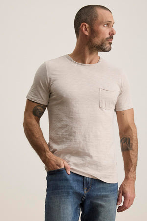 A man with short hair and a beard is wearing the CHAD TEE from Velvet by Graham & Spencer, made of soft Peruvian cotton in light beige, paired with blue jeans. Featuring a crew neckline and subtle chest pocket, he stands tattooed against a plain background looking to his right.