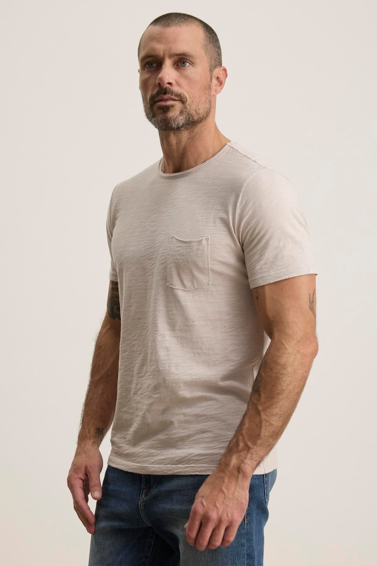   A man with a beard wears the CHAD TEE by Velvet by Graham & Spencer, a soft Peruvian cotton, light-colored t-shirt featuring a crew neckline and subtle chest pocket, paired with jeans as he stands against a plain background. 