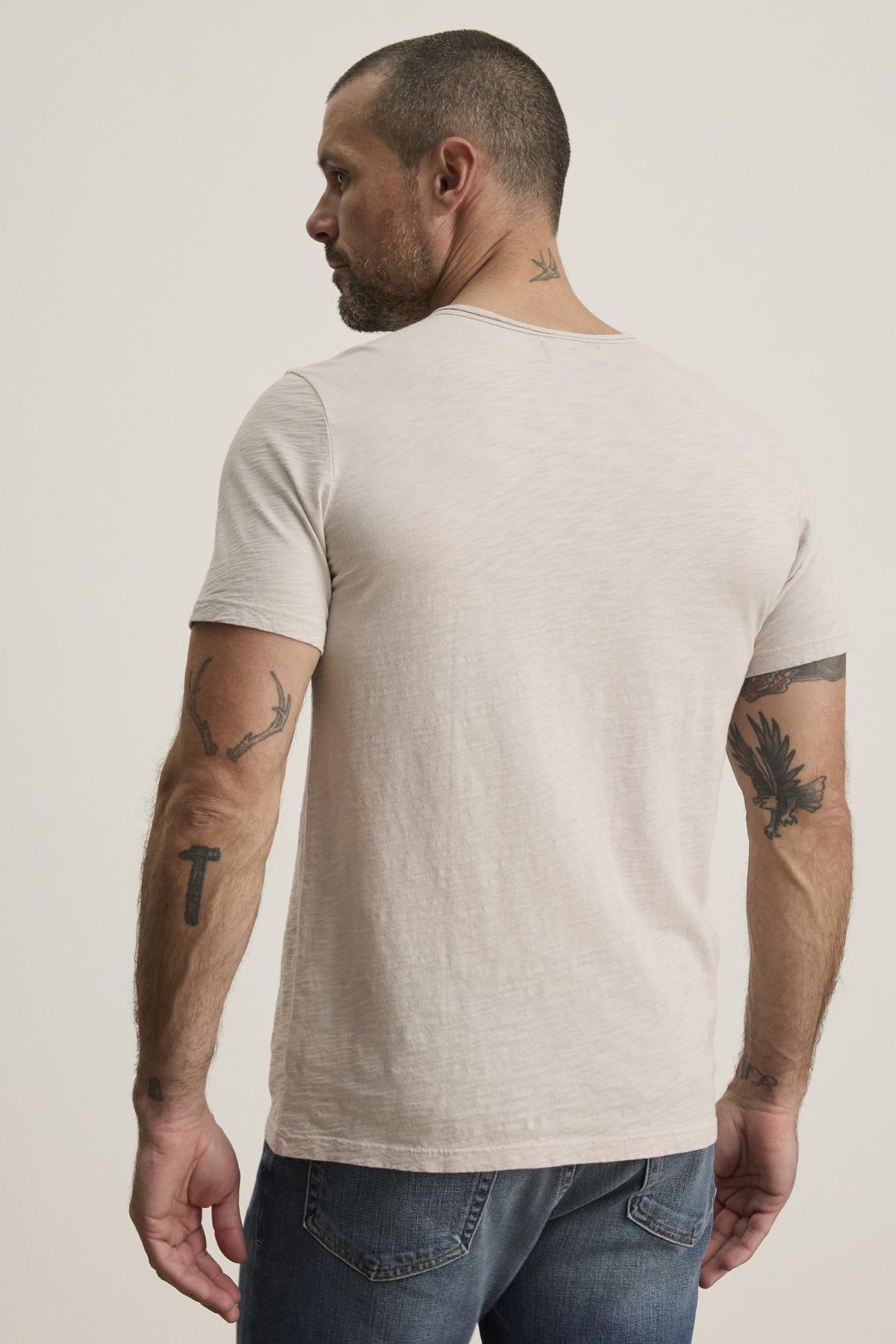   A tattooed man is wearing a CHAD TEE by Velvet by Graham & Spencer in light gray Peruvian cotton with a chest pocket, paired with blue jeans. He stands against a plain background, gazing to the side. 